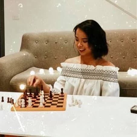 Nguyen Tra My smiling over a chess board on a sofa at home, in a white off-the-shoulder top