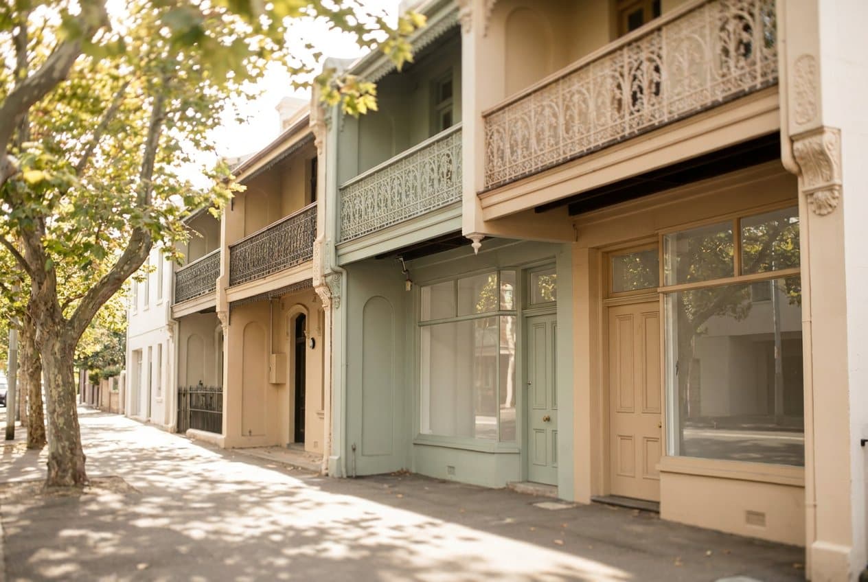 A street of two-storey Victorian terraces with iron lace balconies above small shopfronts, plane trees in afternoon light