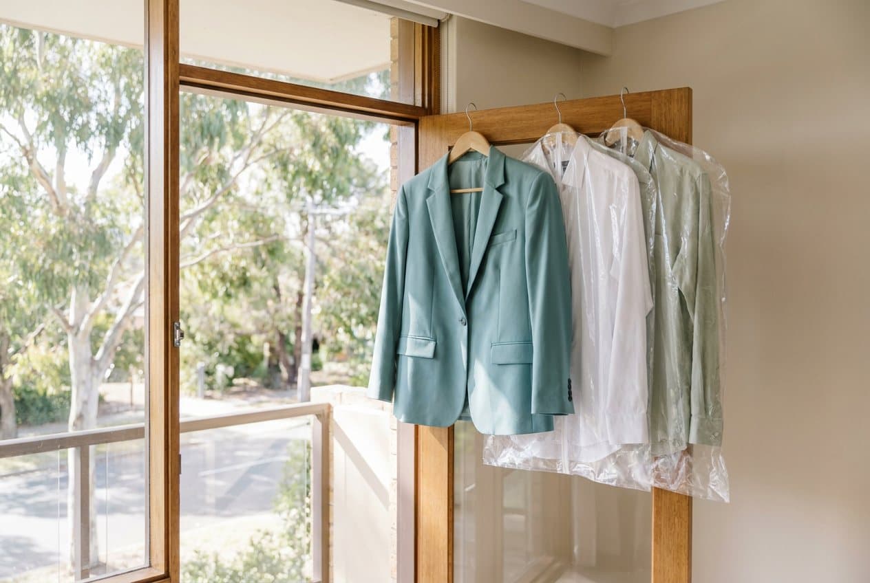 A pressed blazer and two business shirts in clear covers hanging by a bright apartment balcony door